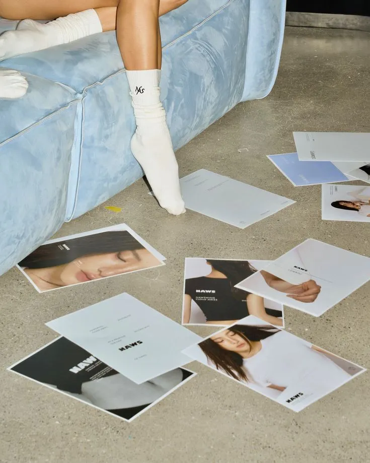 Printed layouts spread across a concrete floor beside a pale blue sofa